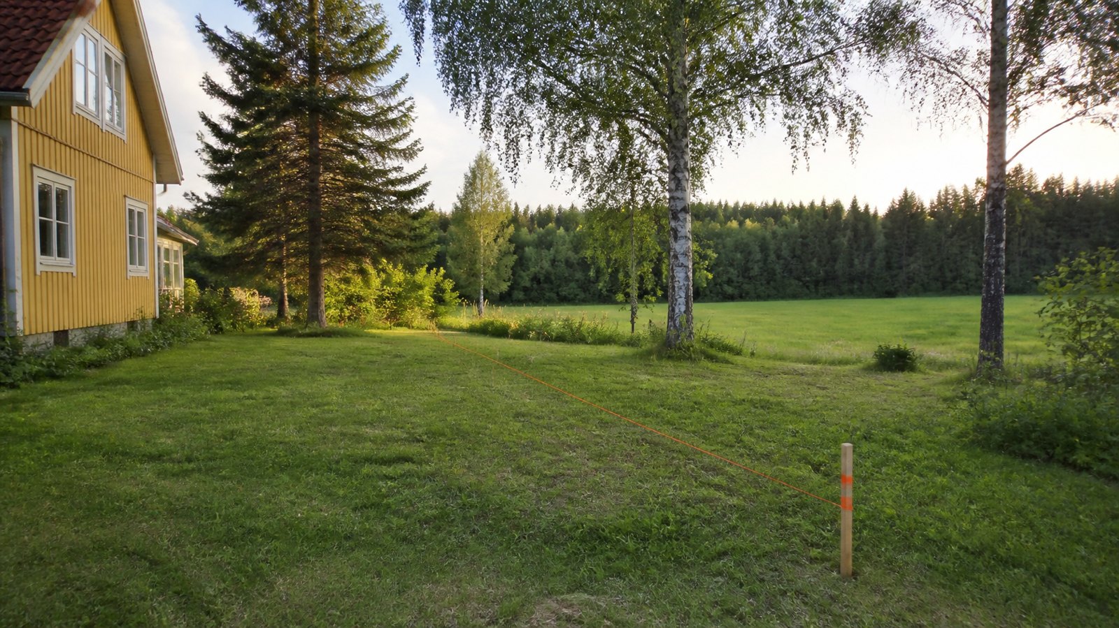 Gul trävilla och gräsmatta där en ny tomtgräns är markerad med käpp och orange snöre bland björkar i Sverige