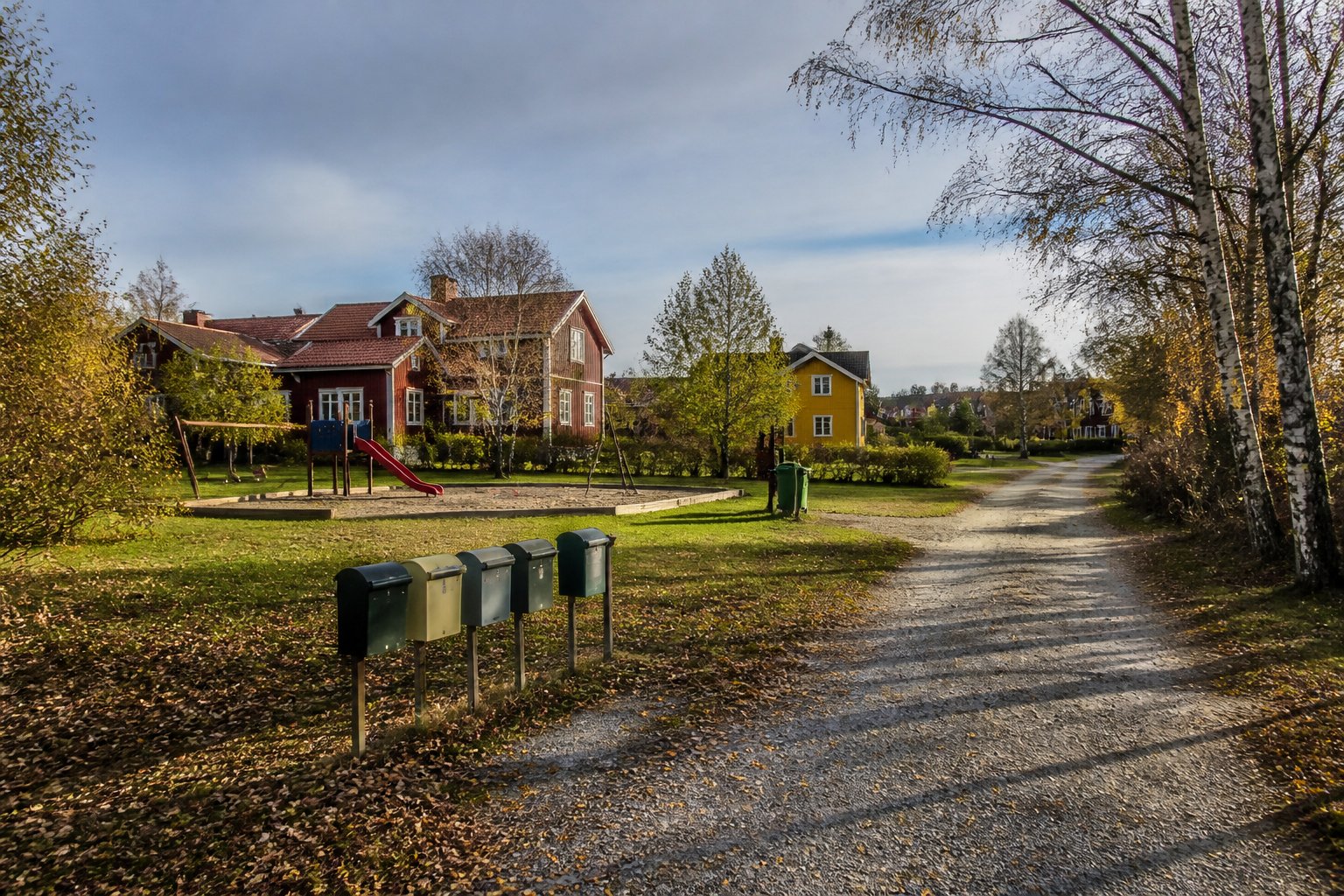 Gemensam grusväg, lekplats och brevlådor i ett svenskt villaområde med röda och gula trähus – gemensamhetsanläggning
