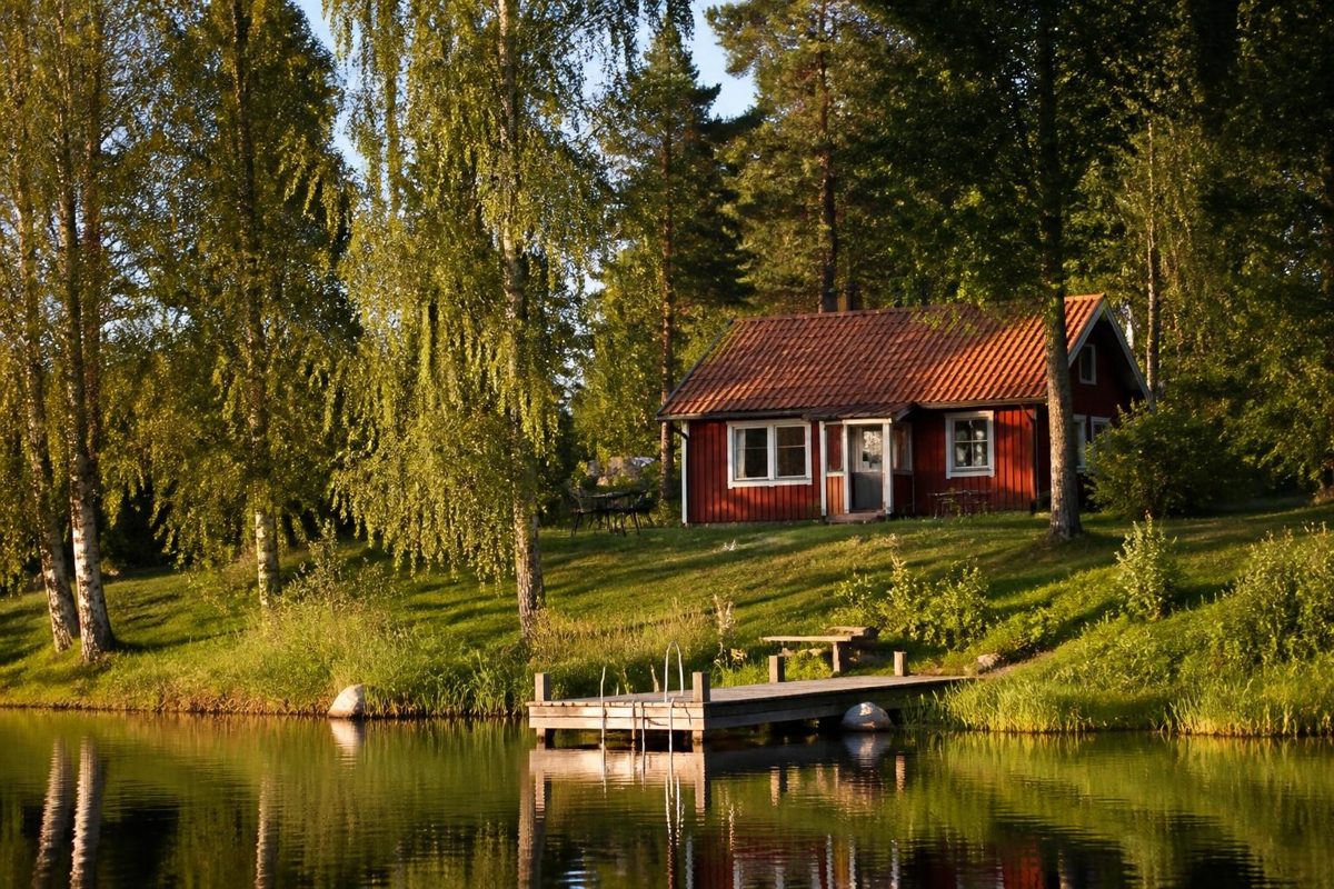 Rött svenskt trähus med vita knutar på en gräsbacke vid en sjö med brygga och björkar